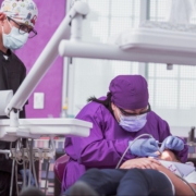 A female dentist wearing white scrubs, blue gloves, and a mask performs a dental procedure on a woman with pink hair while a female dental hygienist is helping.