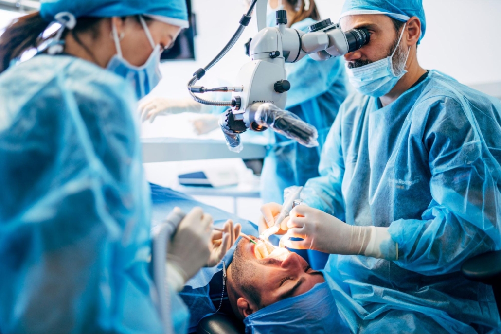 Two dentists wearing blue PPE performing an oral surgery on a male patient, with a female dental assistant in the background.