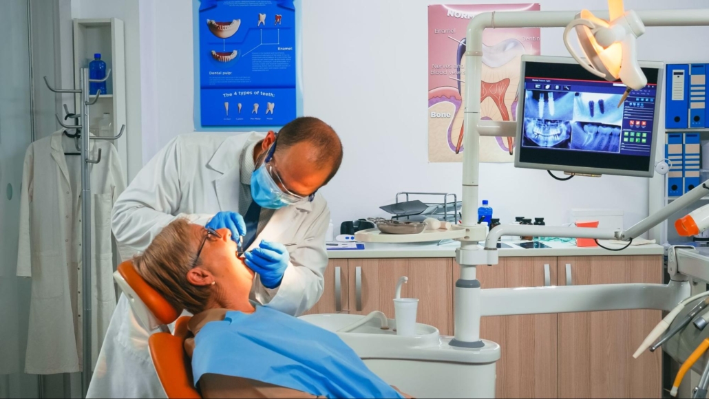 A male dentist wearing a white lab coat, a mask, and blue gloves is doing a dental exam of a woman on a dentist’s chair.
