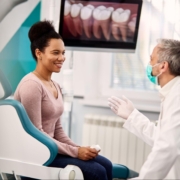A female patient in a dental office having a conversation with the dentist while an x-ray is on a screen behind them.
