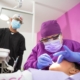 A female dentist in purple scrubs performing a dental procedure on a patient with her assistant.