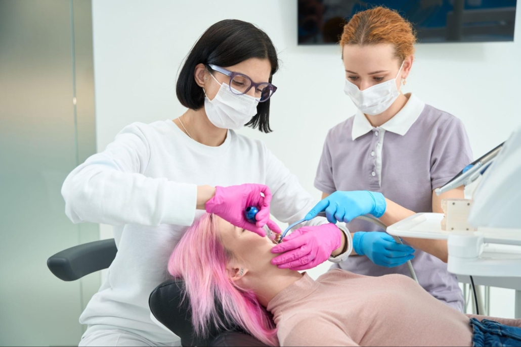A female dentist wearing white scrubs, blue gloves, and a mask performs a dental procedure on a woman with pink hair while a female dental hygienist is helping.