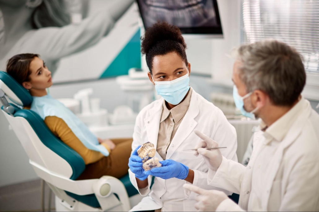 Two dentists in white coats and masks communicate with each other while a female patient is in the background looking at them while in the dental chair.