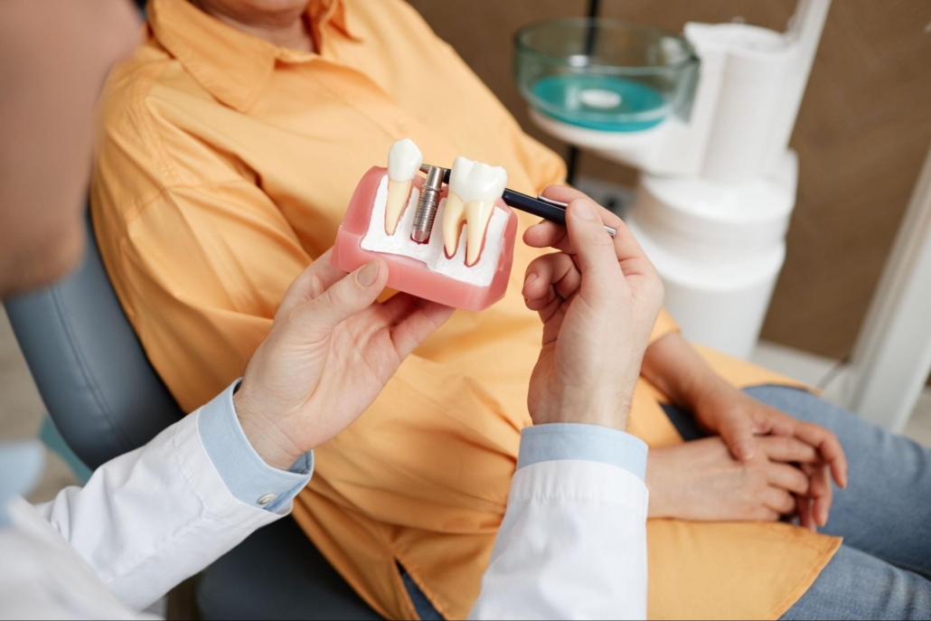 Patient in a chair next to a dentist holding a model of dental implants. 