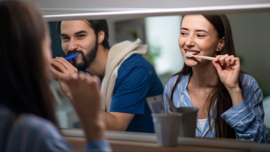 A man with brown hair and a beard wearing a blue shirt and a woman with long brown hair wearing pajamas smiling at each other while brushing their teeth in a mirror in a bathroom.