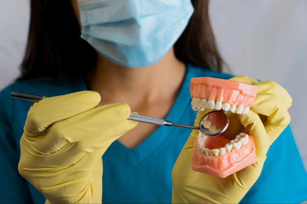 A female dental hygienist wearing blue scrubs and a mask posing while holding a model of prosthetic teeth and a dental mirror. 
